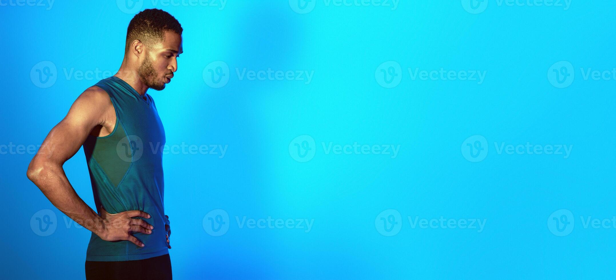 Black Man In Sportswear Doing Breathing Exercise Getting Ready For Workout Standing On Blue Studio Background. Empty Space, Neon Lights photo