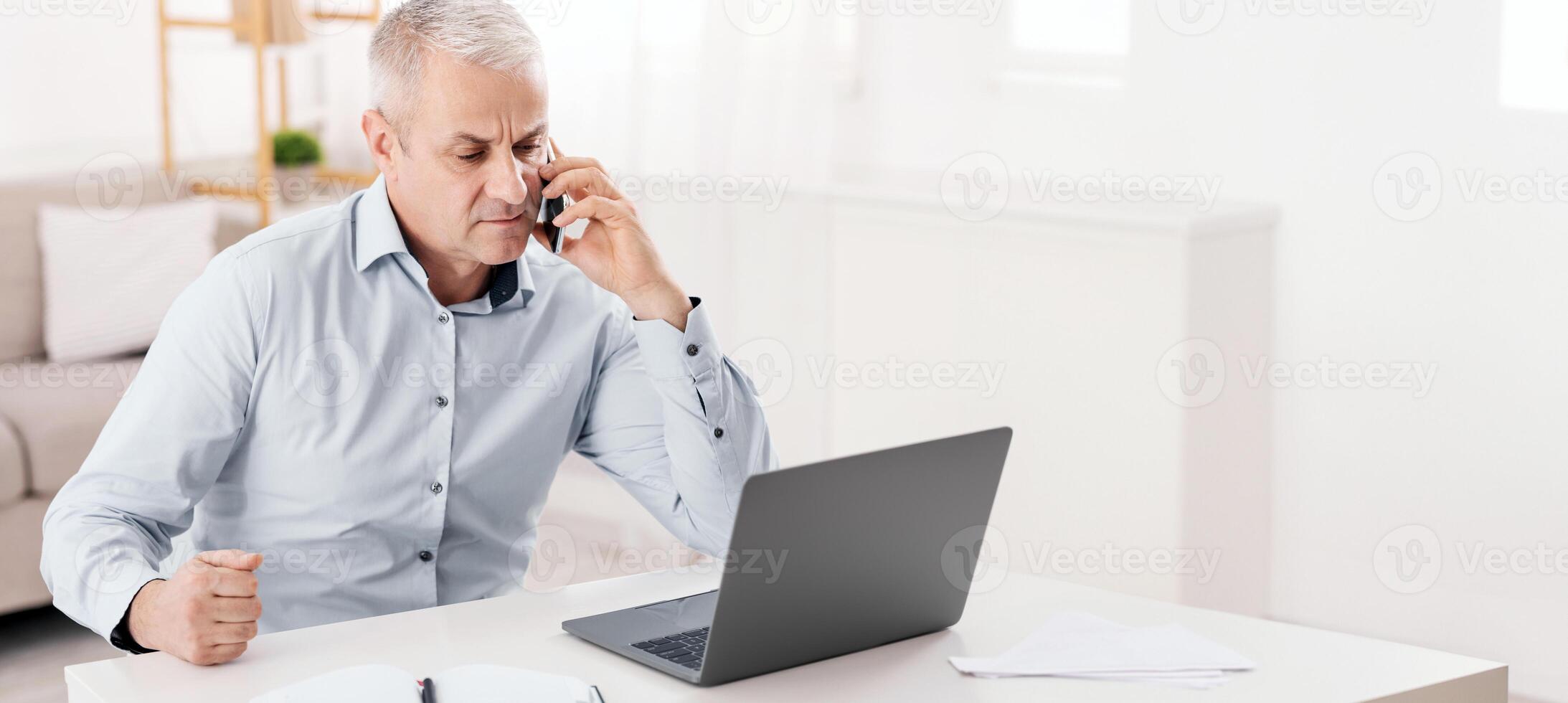 Business Calls Concept. Concerned Senior Man Talking On Mobile Phone Sitting At Desk, Looking At Laptop, Empty Space photo