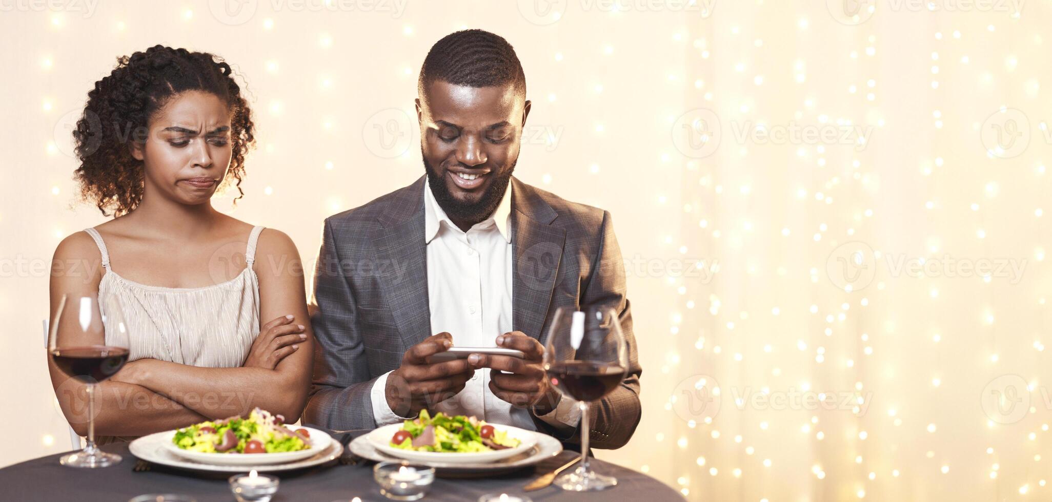 Annoyed afro woman looking at her man using phone while having date in restaurant, conflicts in couple concept photo