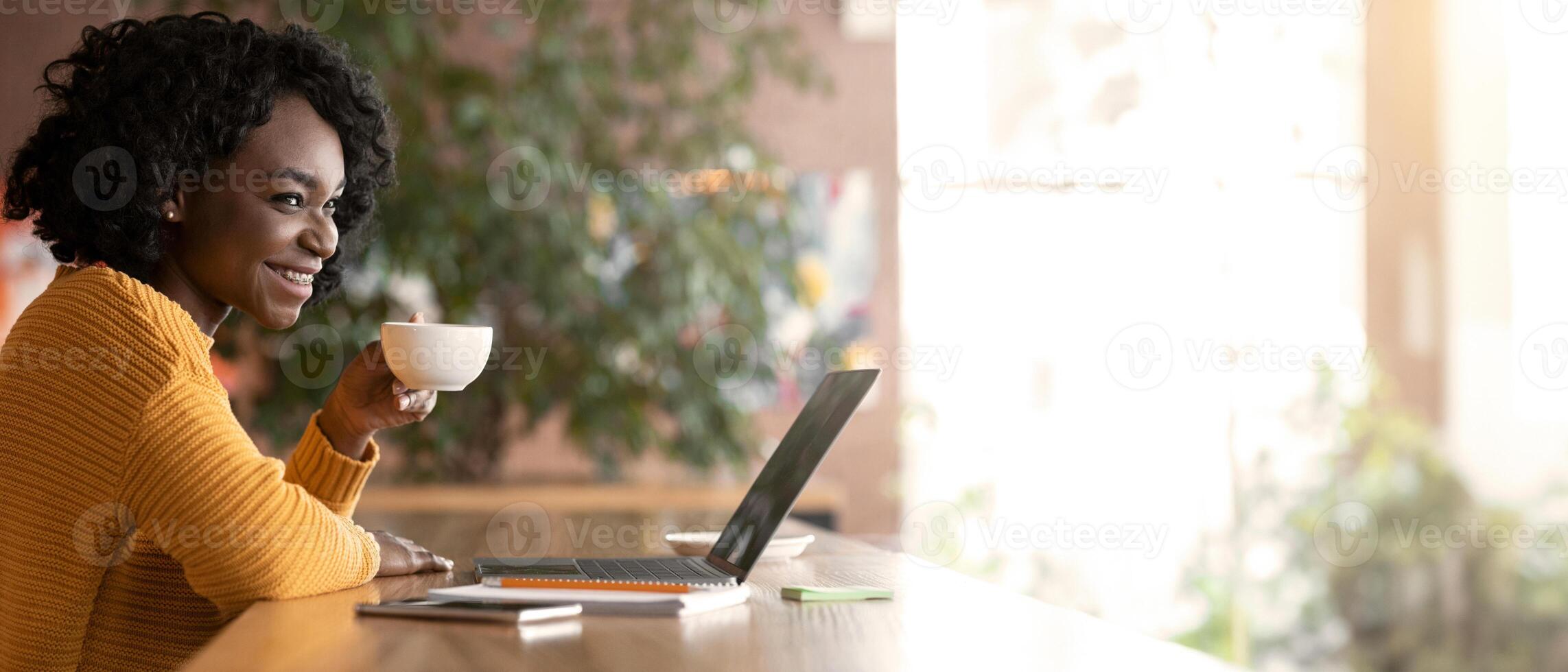 Happy african girl drinking tea while working with laptop at cafe, empty space photo