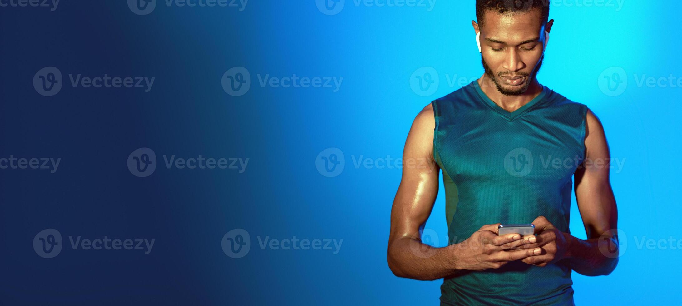 Fitness Application. Sporty Guy Using Smartphone Wearing Earphones Before Workout Standing On Blue Studio Background. Panorama photo