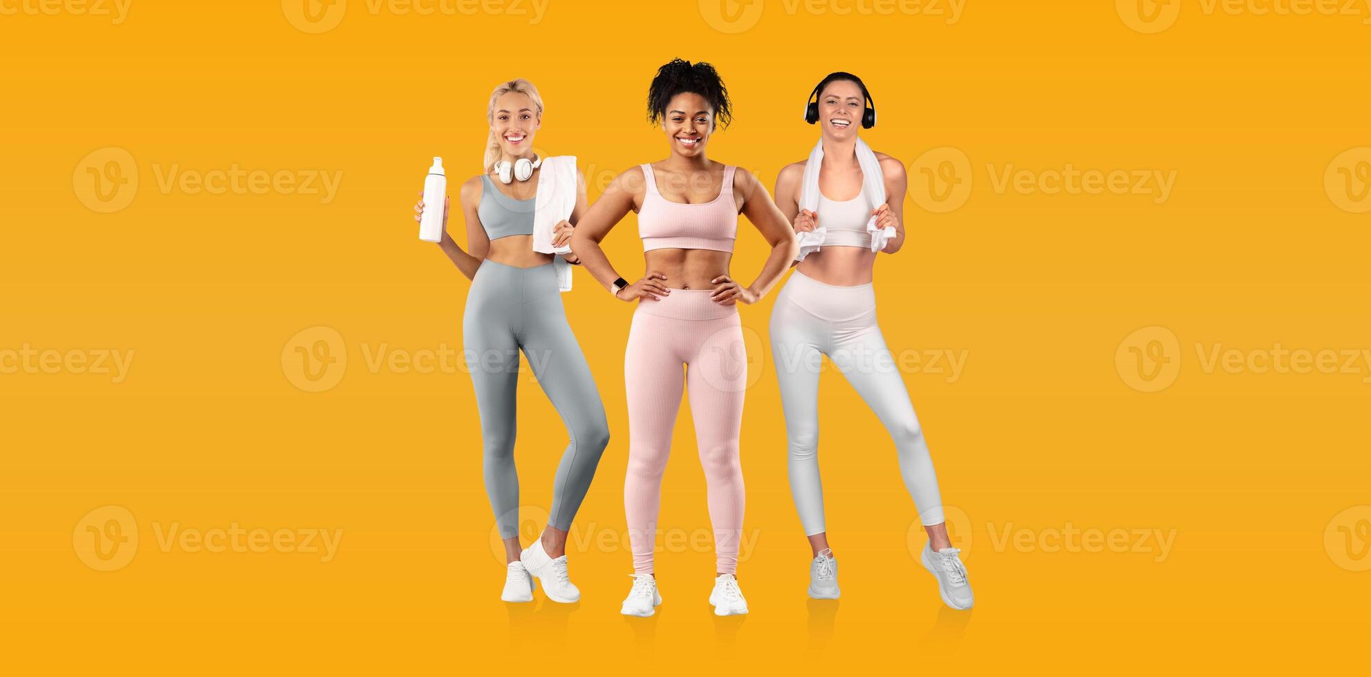 Three women dressed in colorful activewear stand together, each holding water bottles, smiling brightly. The vibrant yellow backdrop enhances their energetic vibe. photo