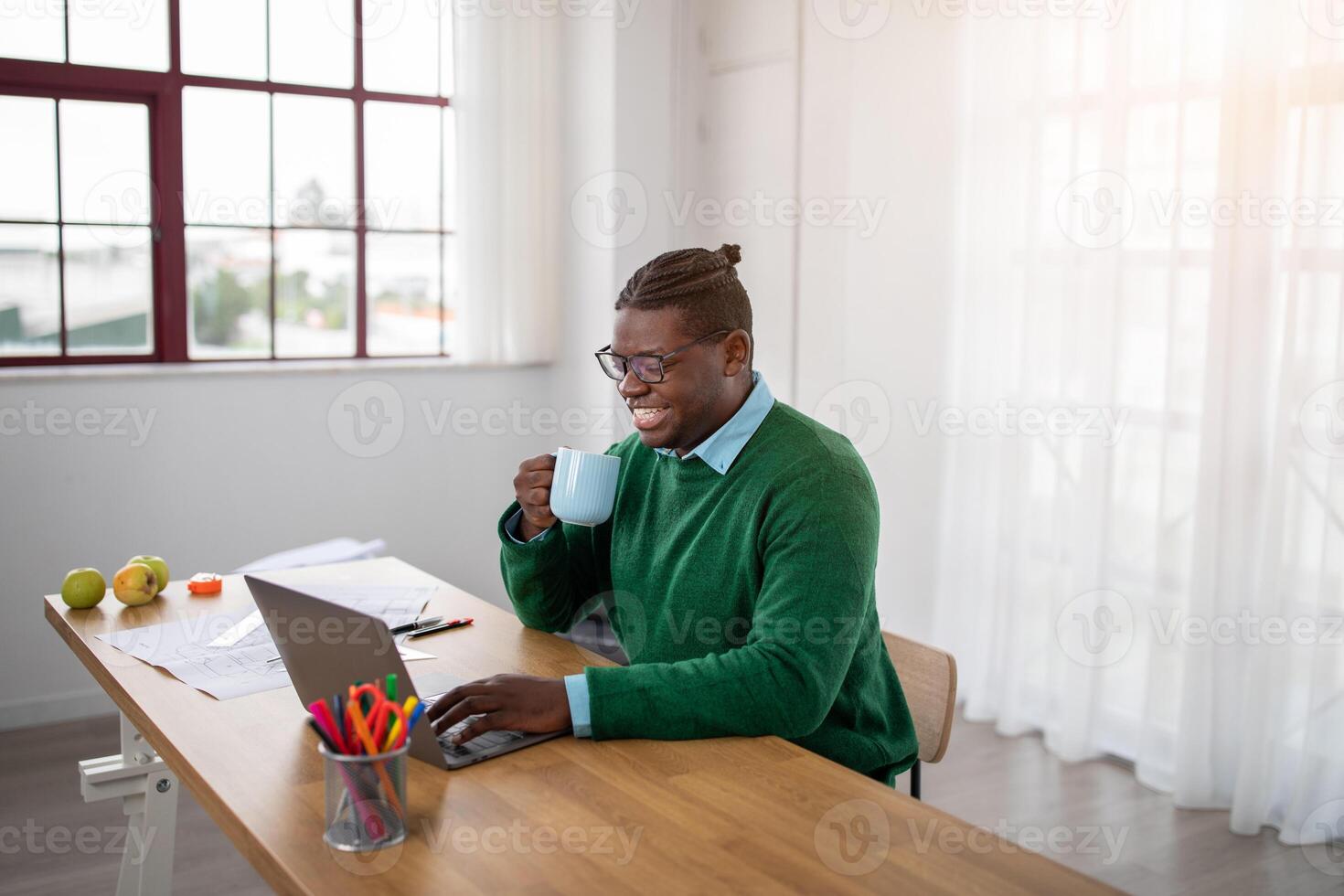 A cheerful black man in a green sweater sits at a wooden desk, sipping coffee from a mug while working on his laptop. The sun streams through the windows, creating a warm atmosphere. photo