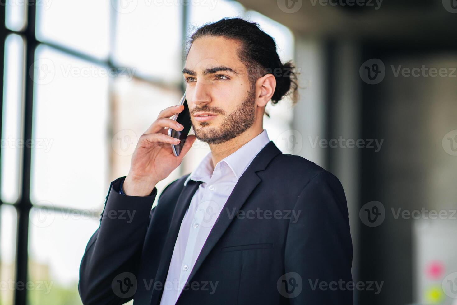 A business professional wearing a suit is talking on a mobile phone inside a modern office setting. Sunlight streams through large windows, creating a dynamic atmosphere. photo