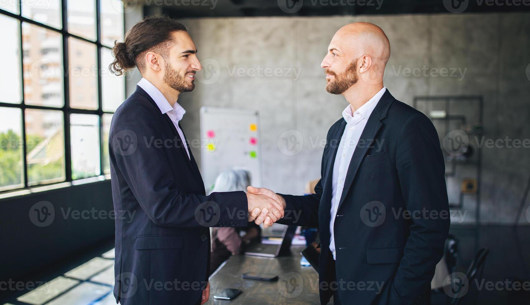 Two men in suits greet each other with a handshake in a contemporary office. A group of colleagues is engaged in discussion in the background, emphasizing collaboration and professionalism. photo