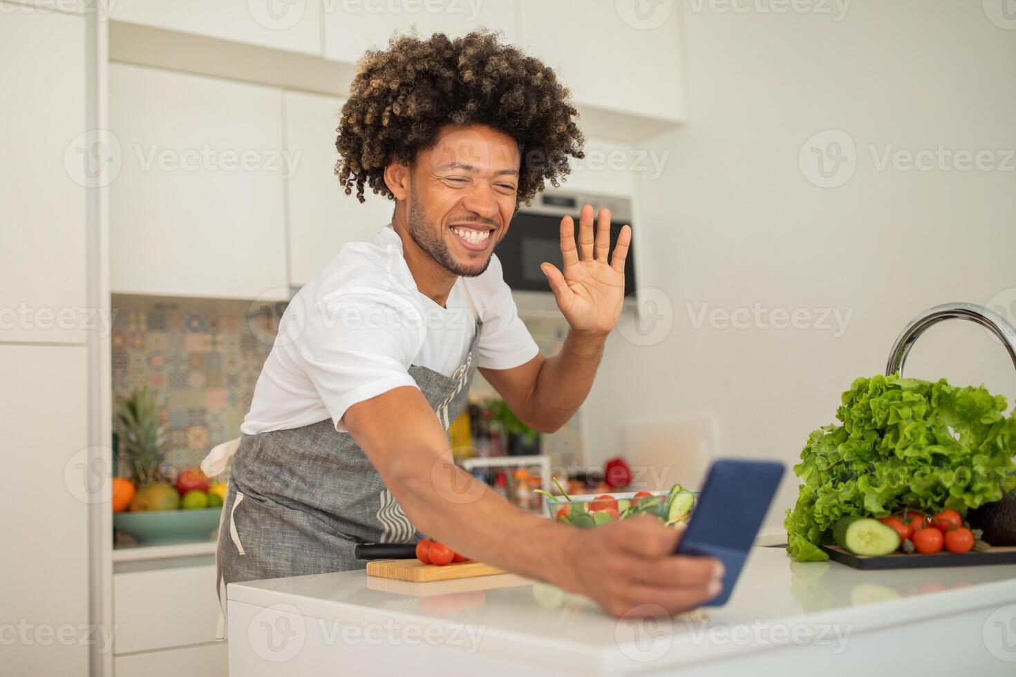 A cheerful man with curly hair is preparing a meal in his bright kitchen, smiling and waving at his phone to connect with friends during a chat. Fresh vegetables are scattered on the counter. photo