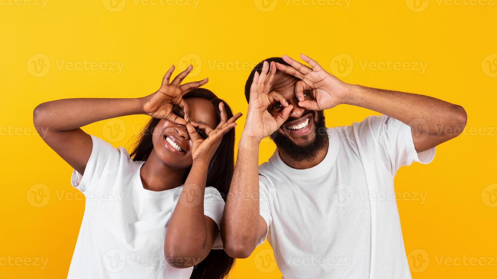Portrait of joyful African American couple smiling, looking at the camera through fingers, doing okay gesture and covering eyes, imitating glasses. Facial expressions and emotions concept photo