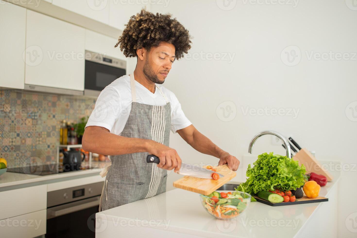 A black man with curly hair is chopping vegetables on a wooden cutting board in a stylish kitchen. Fresh ingredients are neatly arranged, showcasing a healthy cooking environment. photo