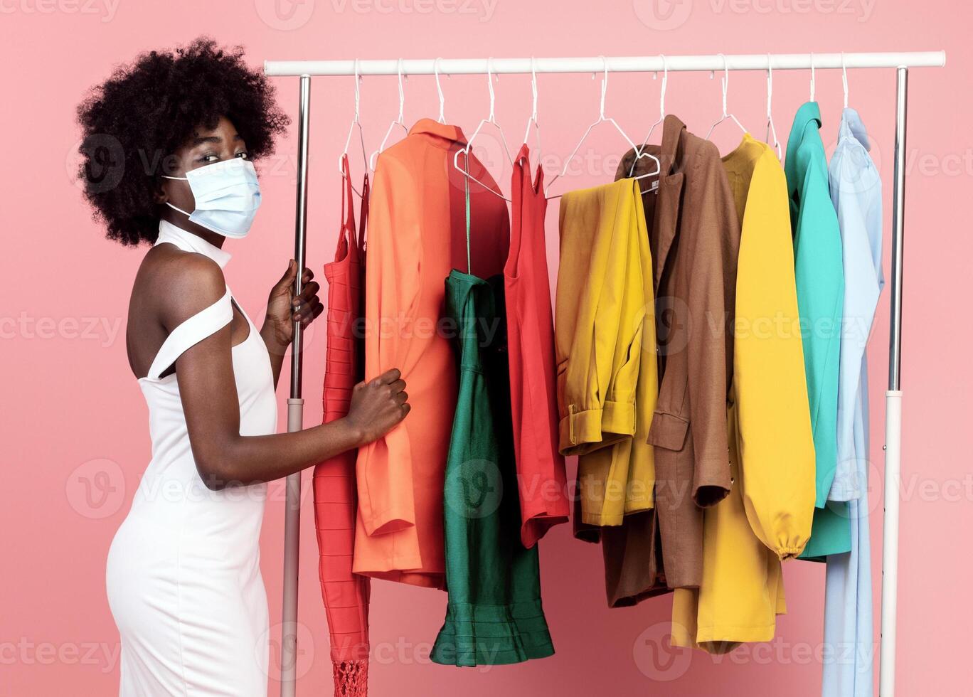 Safe Shopping. Black Lady Wearing Protective Face Mask Choosing Clothes Standing Near Clothing Rack Over Pink Studio Background, Looking At Camera. Vertical Shot. Covid-19 Protection For Customer photo