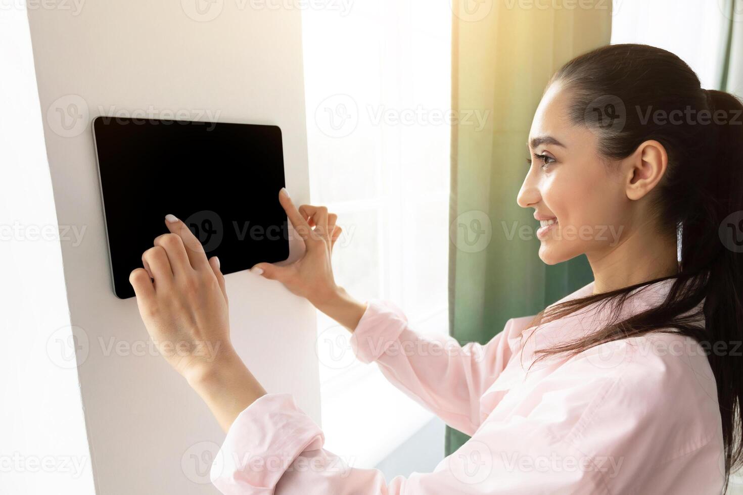 Young woman using thermostat smart home domotic control panel on wall for house temperature. Homeowner touching tablet with empty blank screen to adjust energy saving in living room, iot, mockup photo