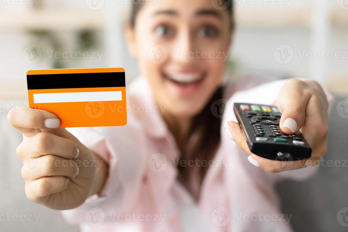 Closeup of amazed excited millennial woman sitting on couch in living room, using tv remote controll and holding yellow golden credit debit card, selective focus on technology, blurred background photo
