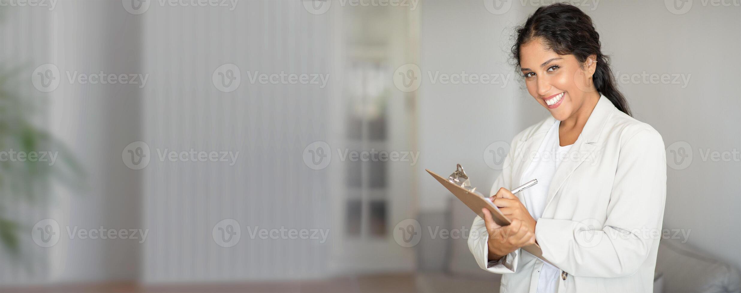 A cheerful healthcare worker stands in a well-lit clinic while holding a clipboard, ready to assist patients with a friendly demeanor and professional appearance. photo
