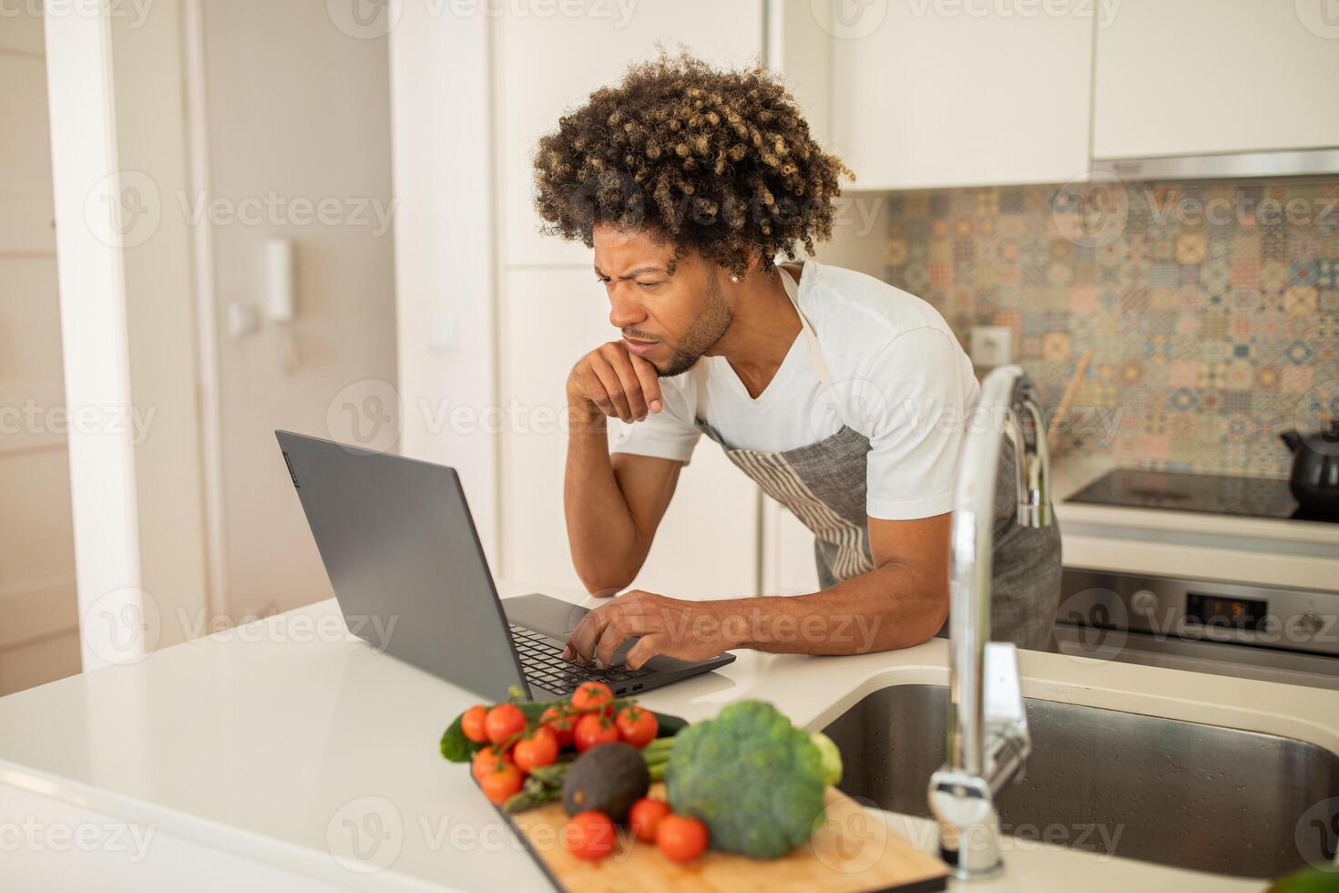 A black man with curly hair is focused on his laptop while surrounded by fresh vegetables in a stylish kitchen. He balances cooking and work, showcasing modern lifestyle choices. photo