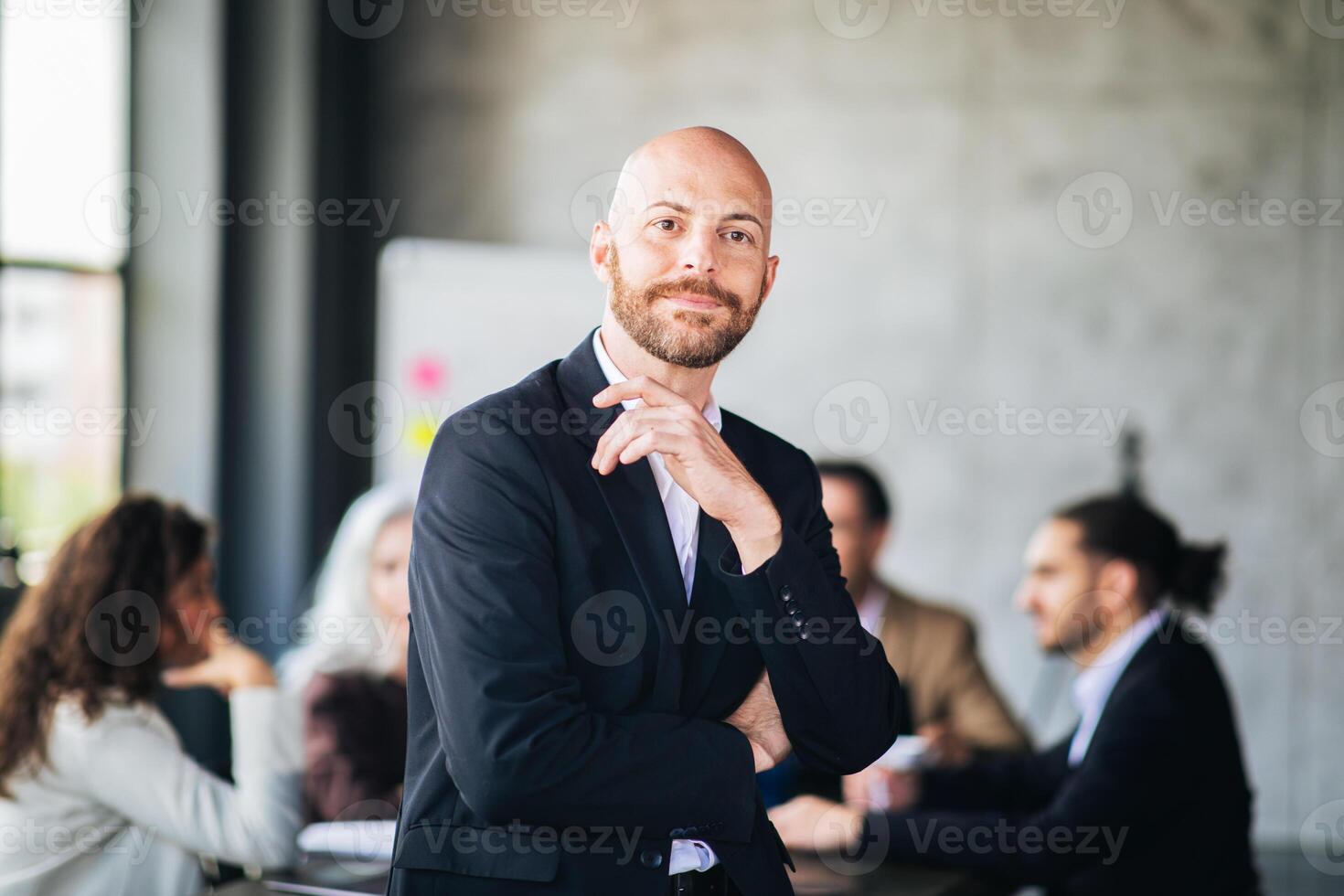 A young man with a beard confidently poses in a stylish suit at a meeting table while colleagues engage in discussion. The modern office has large windows and a contemporary design. photo