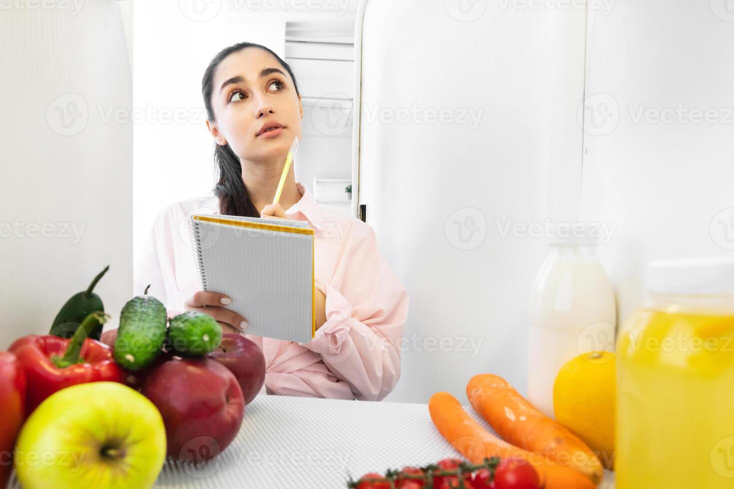 Portrait of young beautiful housewife looking at open refrigerator and thinking, holding notebook with pen taking notes making checklist of necessary food to buy or order, view from inside the fridge photo