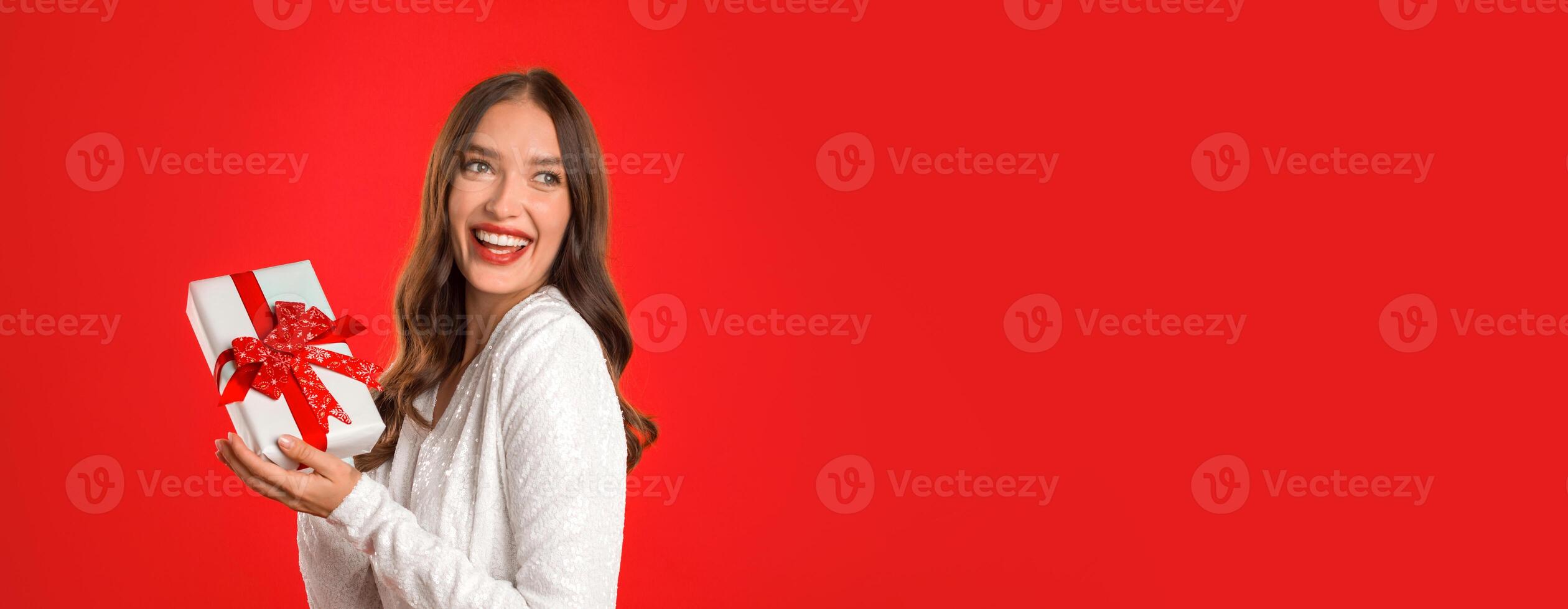 Young woman in evening dress holding gift box and posing on red background. Lady celebrating new year holiday or birthday in elegant clothes holding present, looking aside at free space, panorama photo