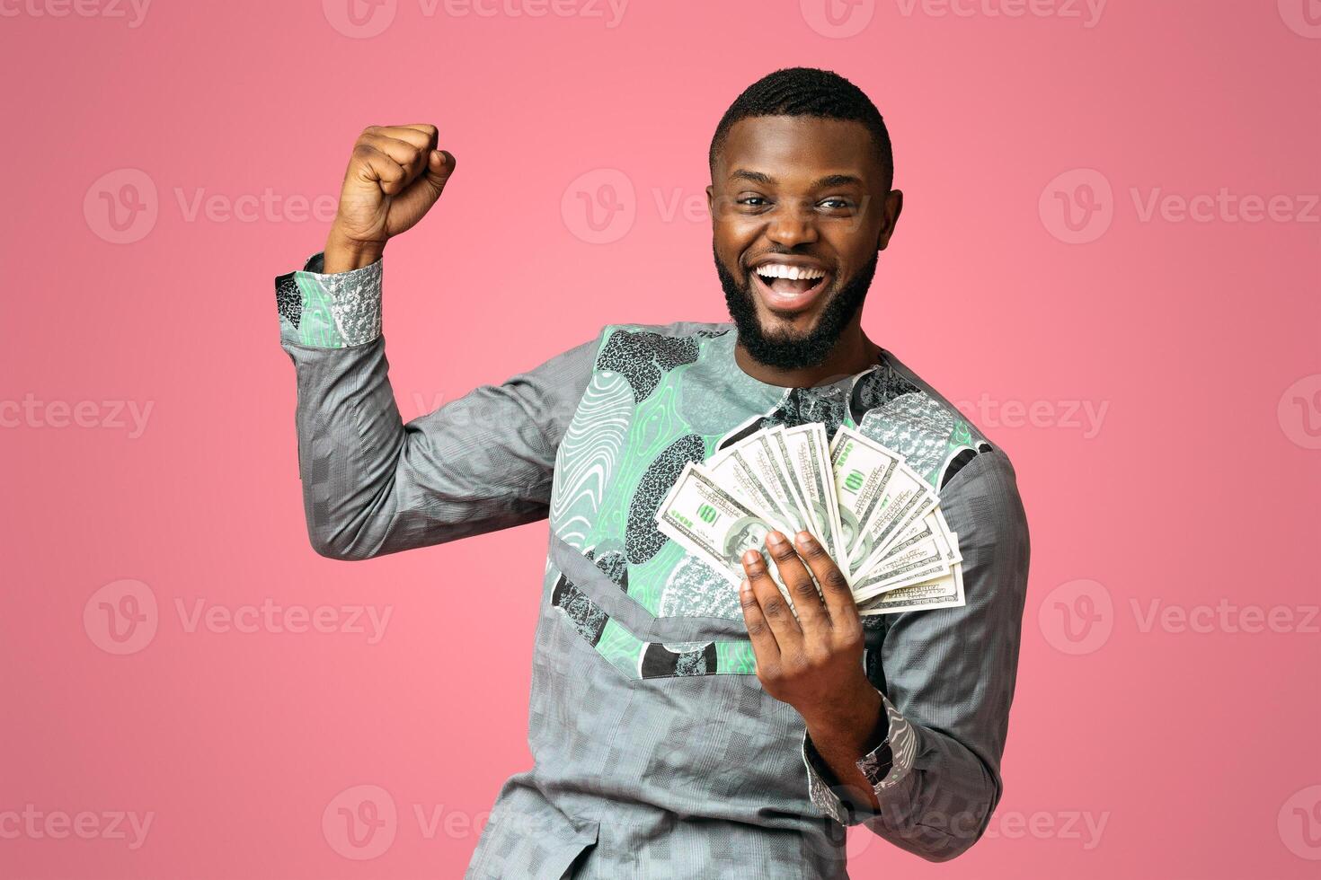 Happy african american young man holding bunch of cash, raising fist up over pink studio background, copy space. Rich and cheerful black guy in traditional costume won prize, gambling concept photo