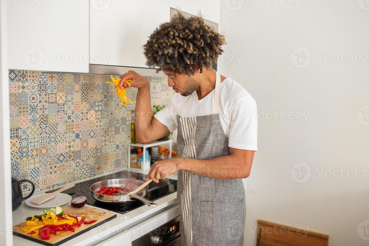 In a well-lit kitchen, a black man with curly hair prepares colorful vegetables, focusing intently while adding fresh ingredients to a pan on the stove. photo