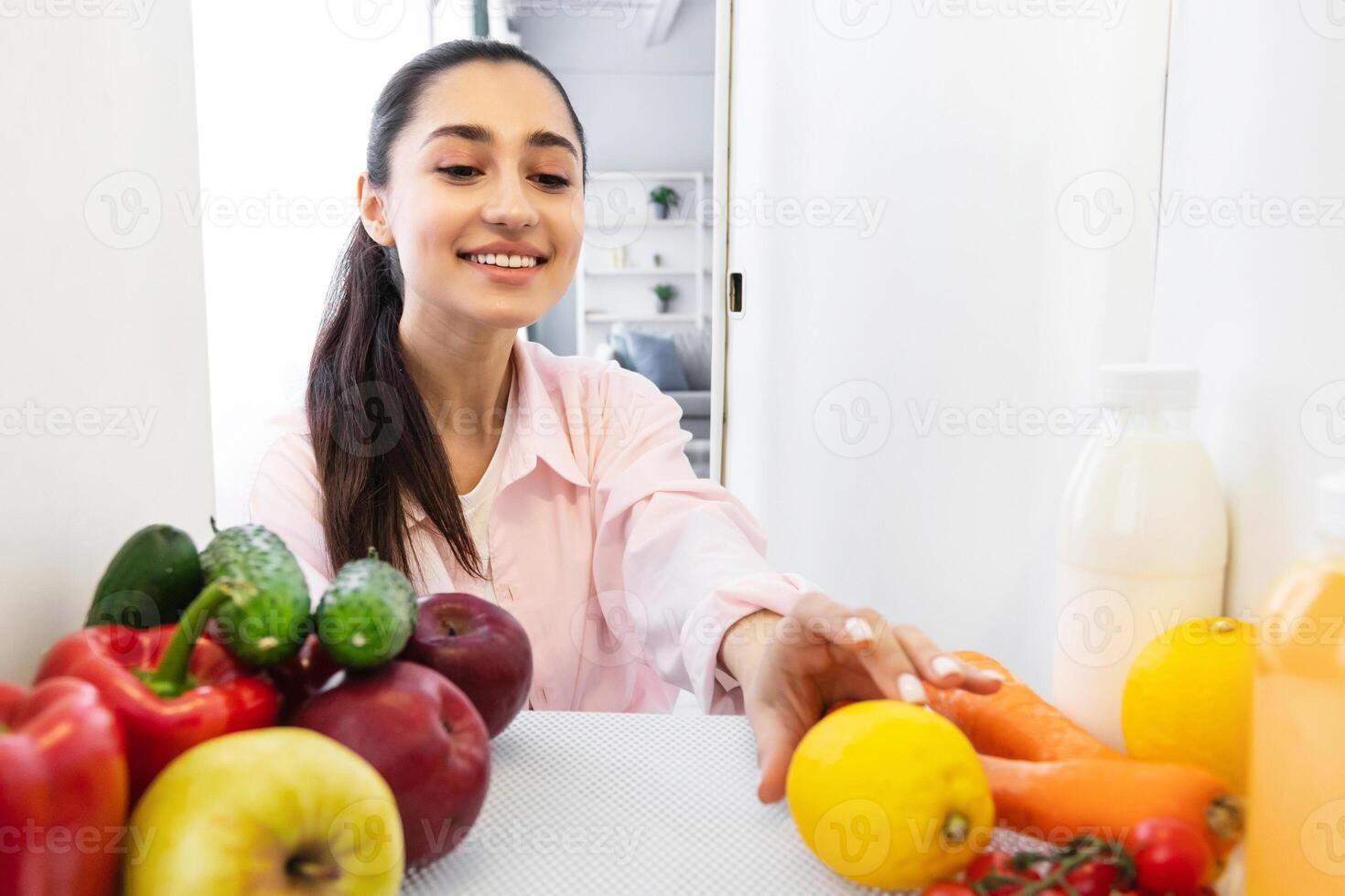 Nutrition Concept. Cheerful millennial housewife opening refrigerator, looking at fruits and vegetables taking fresh citrus, cooking healthy dinner, view from inside the food storage freezer photo