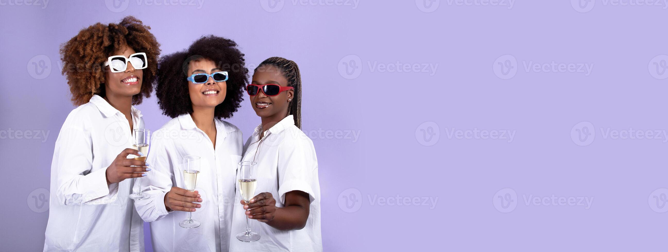 Positive black ladies wearing sunglasses holding glasses with champagne and smiling at camera, isolated on purple background, panorama with copy space photo