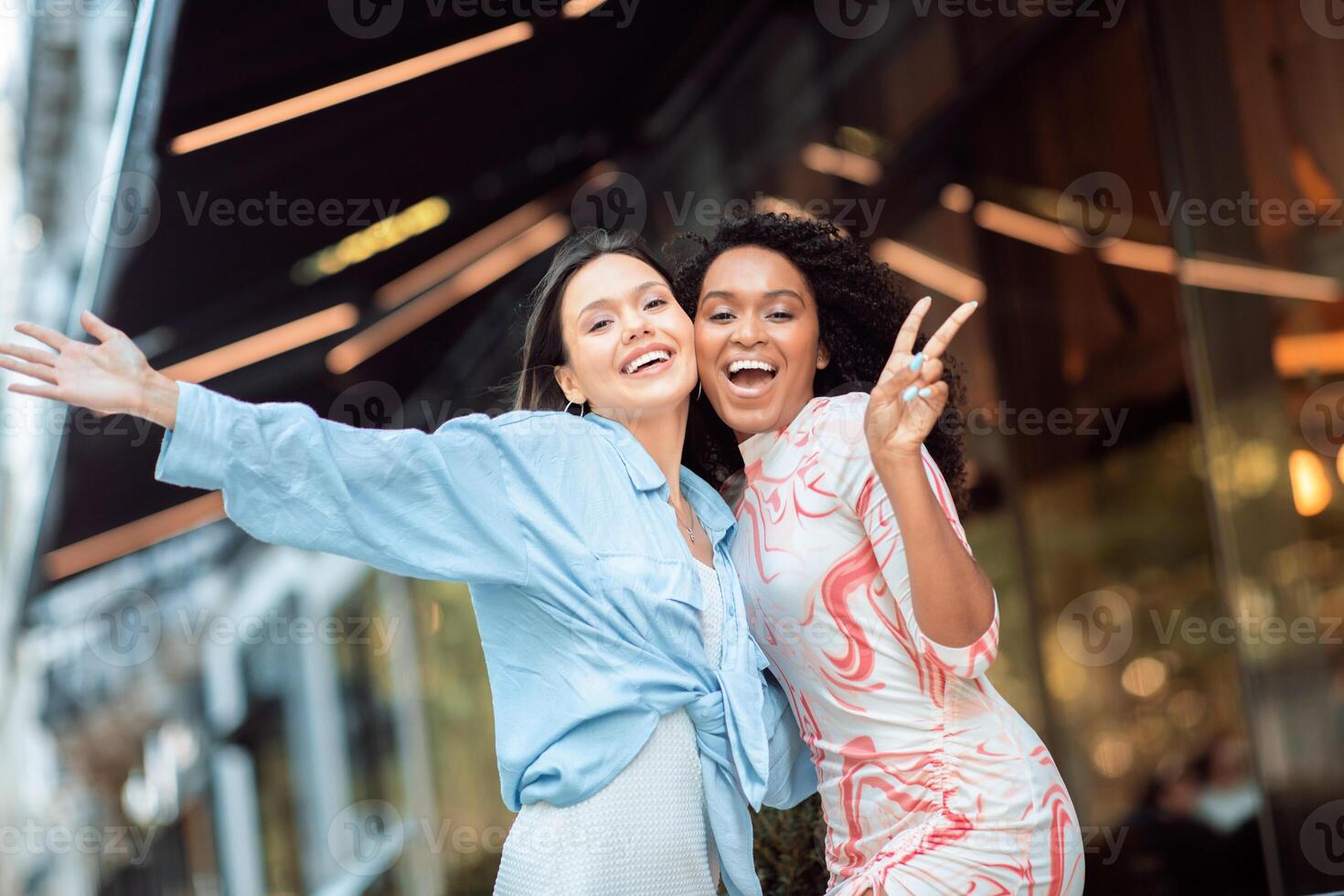 Two women are enjoying a sunny day outside a stylish cafe, smiling and striking a playful pose. They radiate happiness as they celebrate their time together. photo