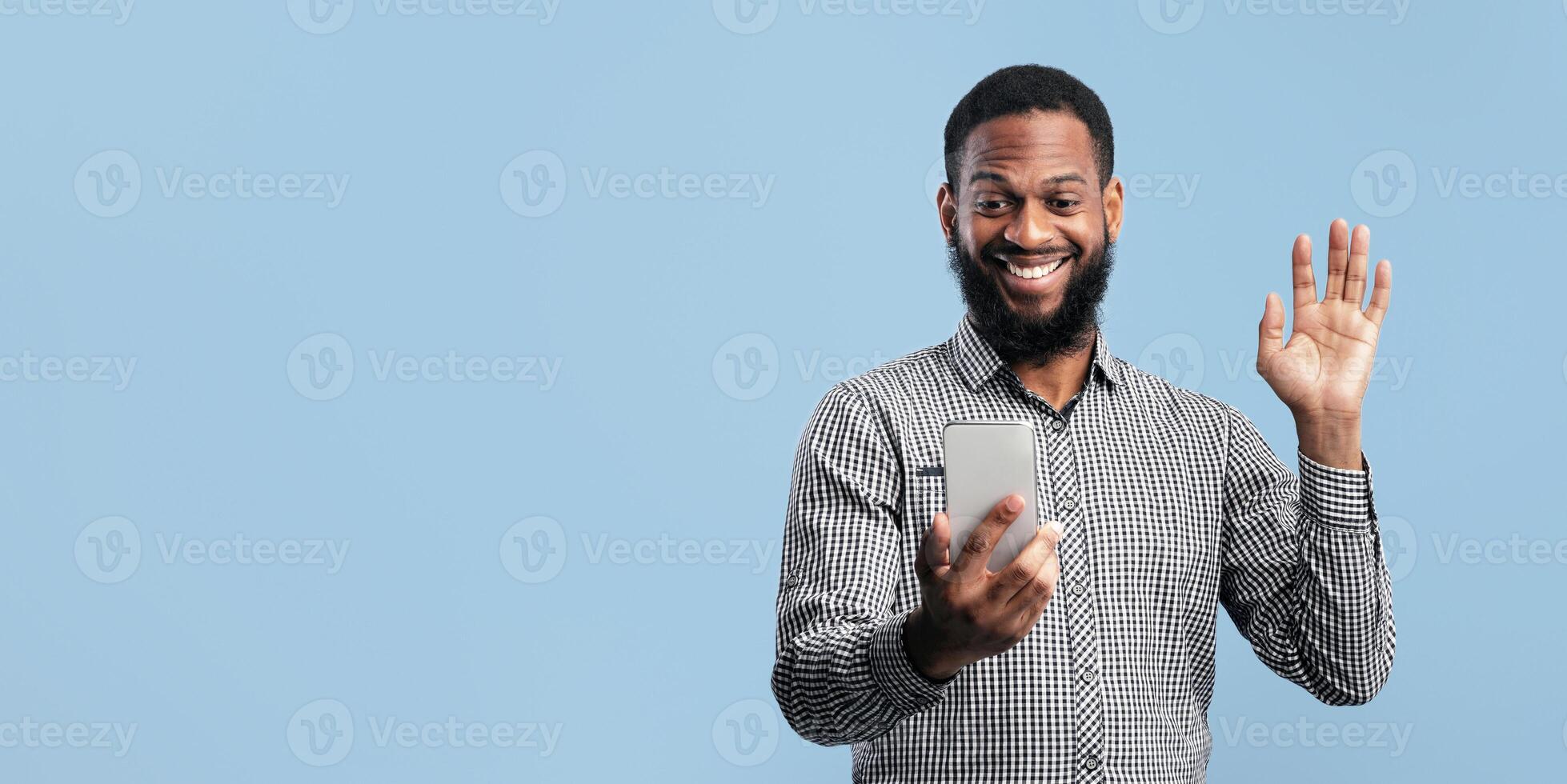 Hello. Front view portrait of excited black guy having call and waving hand at camera, using cell phone, copyspace photo
