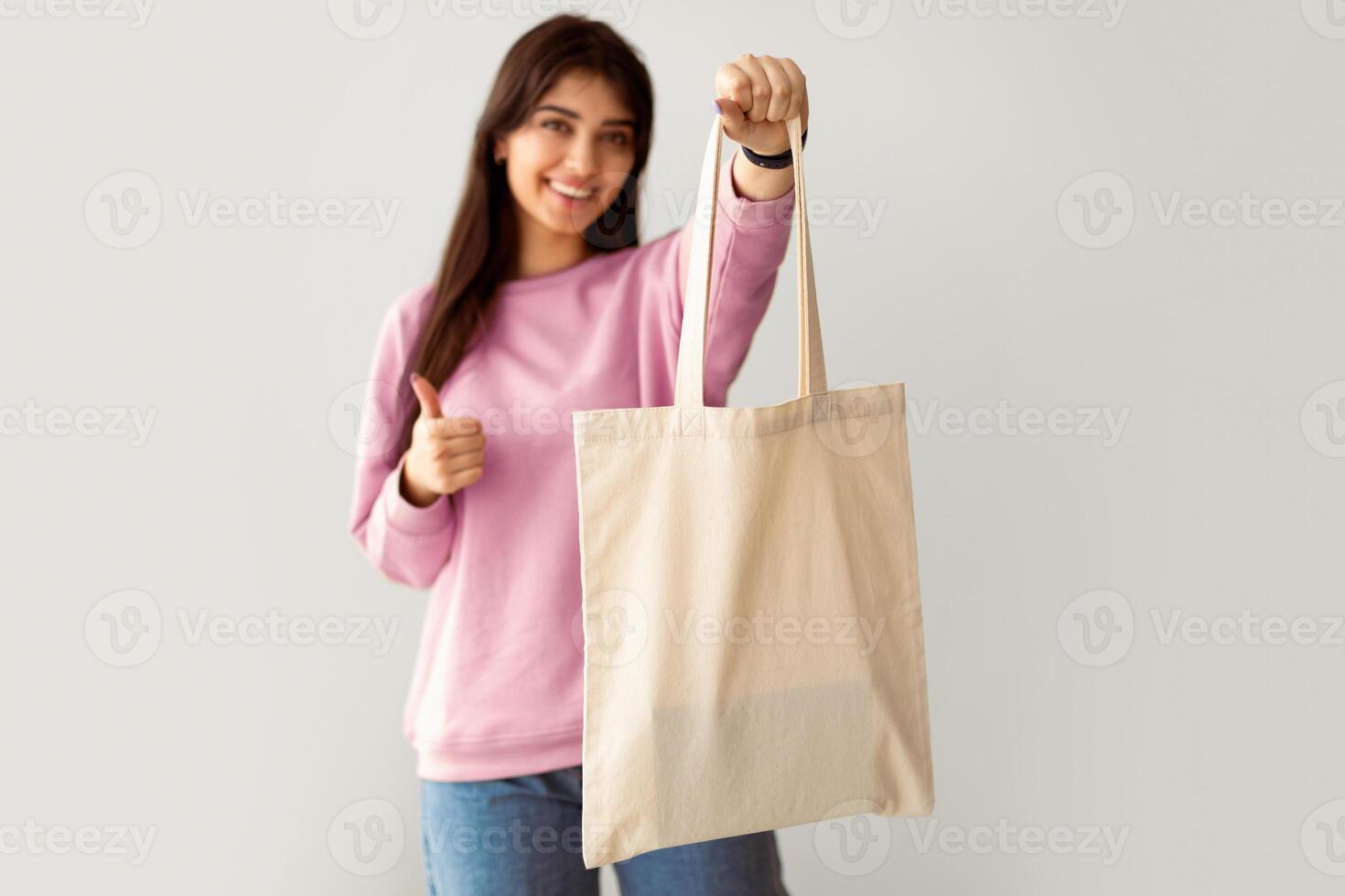 Stop Using Plastic. Cheerful Millennial Woman Showing White Eco Bag To Camera And Doing Thumb Up Sign Gesture. Lady Standing On Studio Background Wall, Holding Flax Shopper Handbag, Selective Focus photo
