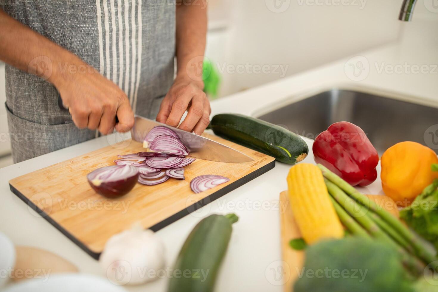 A person skillfully slices red onions on a wooden cutting board in a bright kitchen. Surrounding the board are various fresh vegetables, emphasizing a healthy cooking environment. photo