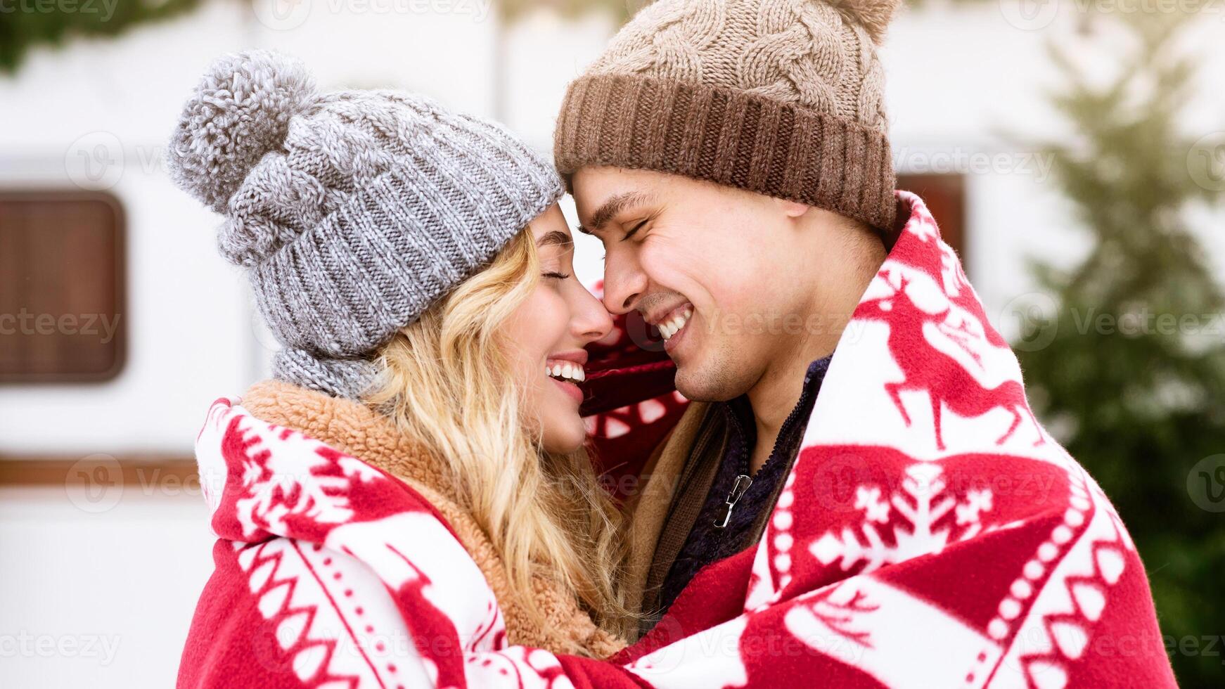 A young couple smiles at each other while wrapped in a red blanket, wearing cozy hats. They enjoy a cold winter morning surrounded by trees, radiating warmth and affection. photo