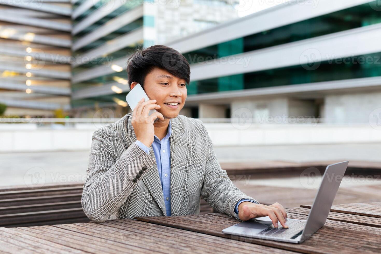A young Asian man in a stylish suit engages in a phone conversation while using a laptop at an outdoor workspace. The modern background features buildings and a sleek environment. photo