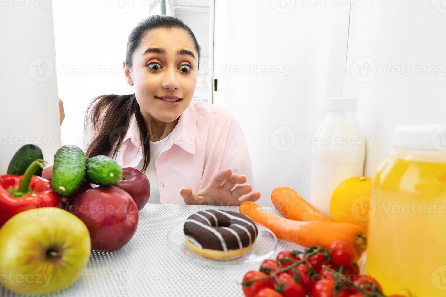 Sugar Addiction Concept. Portrait of funny young lady reaching and trying to grab yummy doughnut pie, view from inside the refrigerator. Sneaky female on a diet at midnight photo