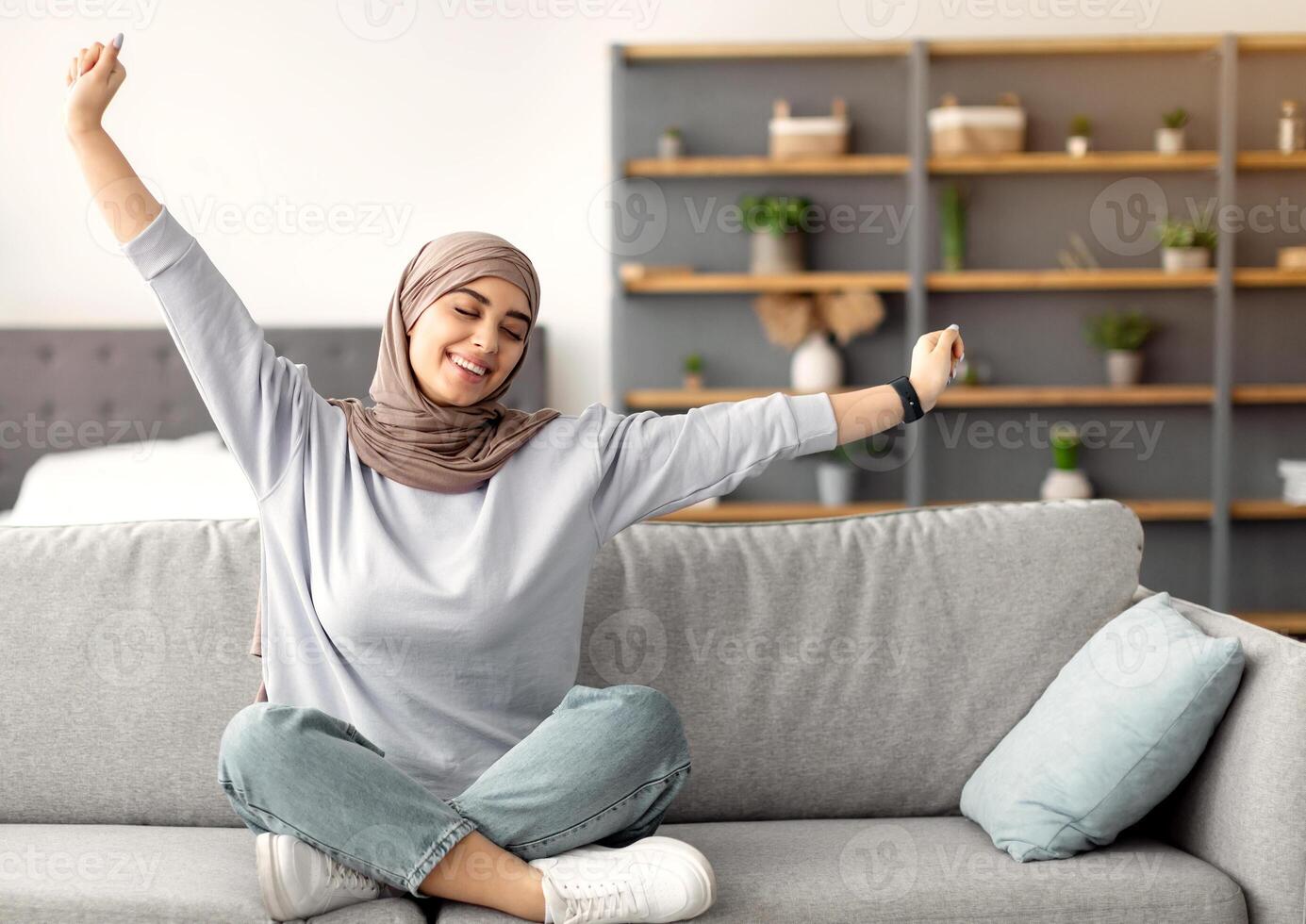 Recreation. Beautiful islamic woman sitting on comfortable couch at home in living room, stretching arms. Happy lady in headscarf relaxing with closed eyes, enjoying free time, taking break from work photo
