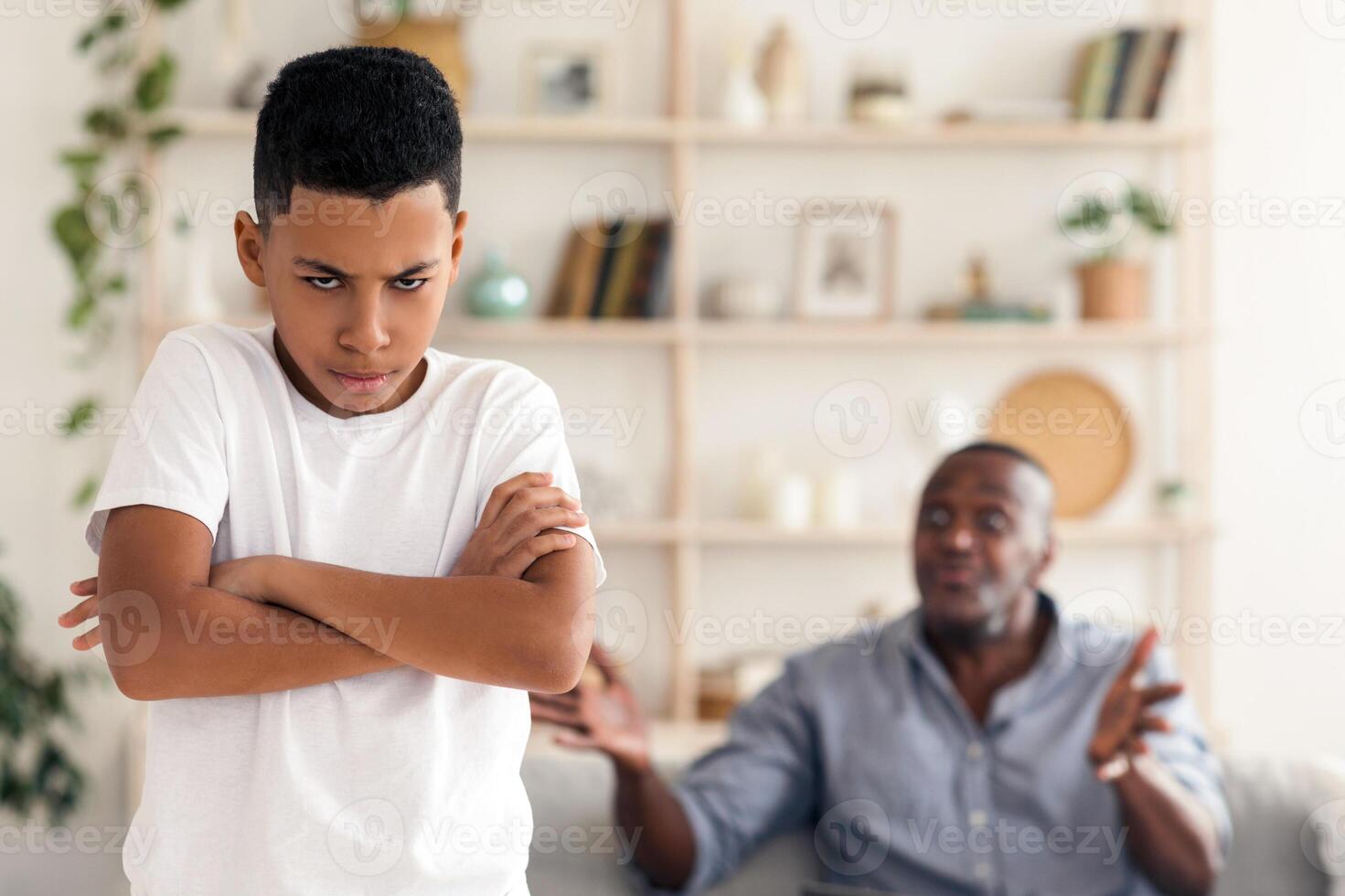 Family problems during quarantine. Angry black grandfather shouting at his grandson at home, focus on kid with folded arms photo