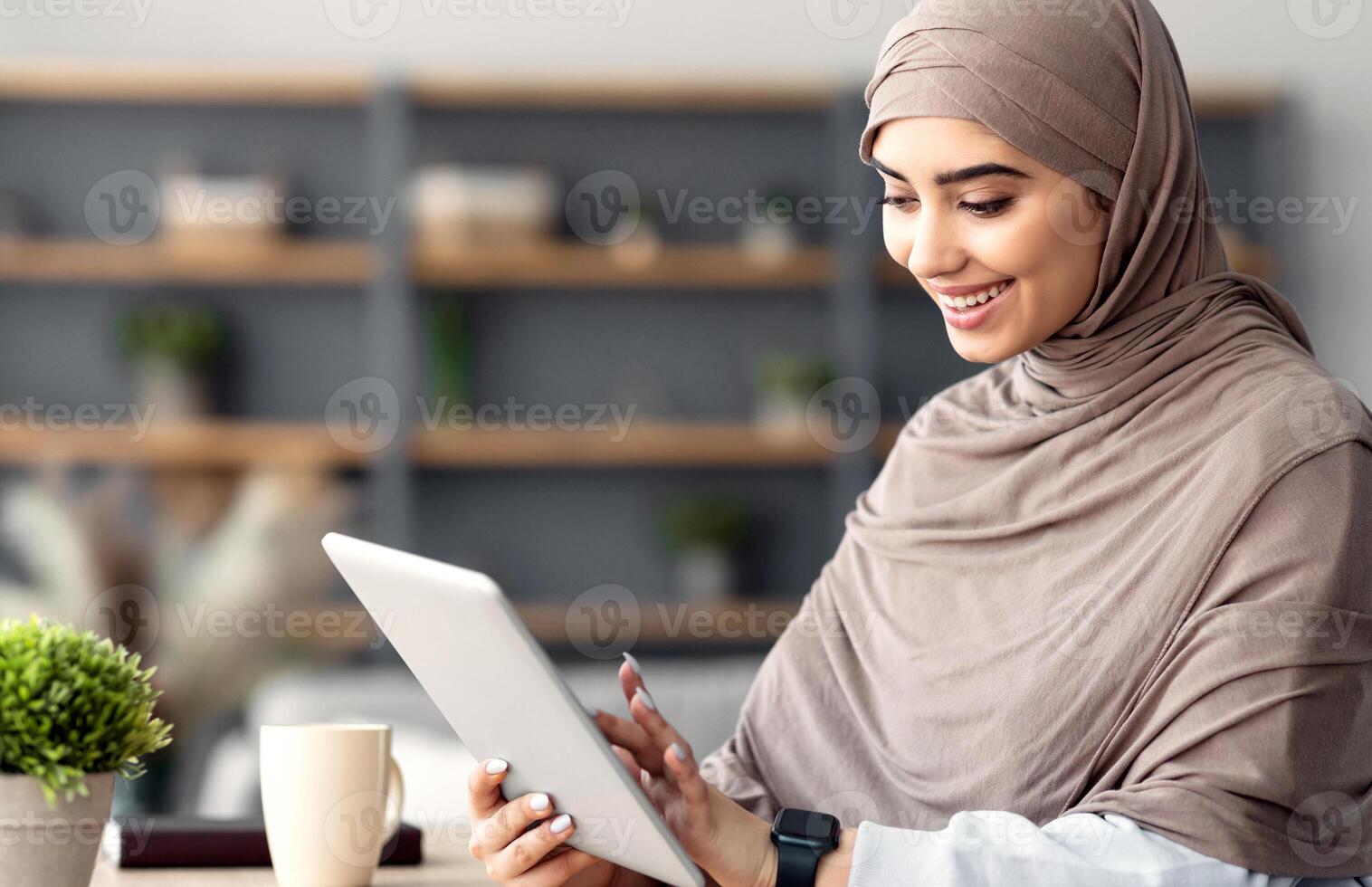 Technology. Millennial Muslim Woman Spending Time With Digital Tablet Sitting At Desk At Home Office, Reading News, Browsing Internet, Watching Videos, Checking Social Networks, Free Copy Space photo