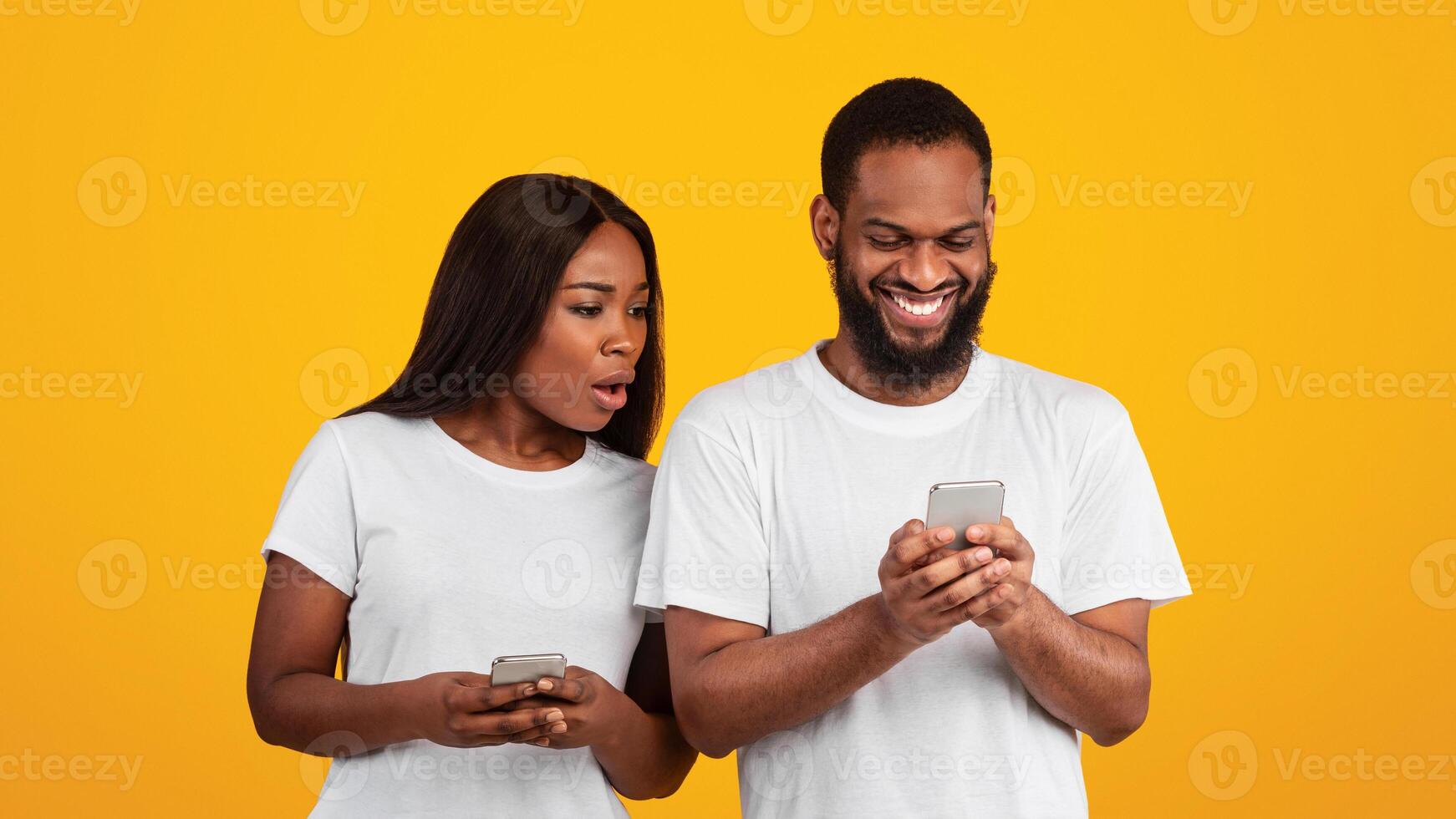 Portrait of shocked African American woman spying on her smiling boyfriend who using mobile phone, texting sms or scrolling social media news feed. Black couple standing over yellow studio background photo