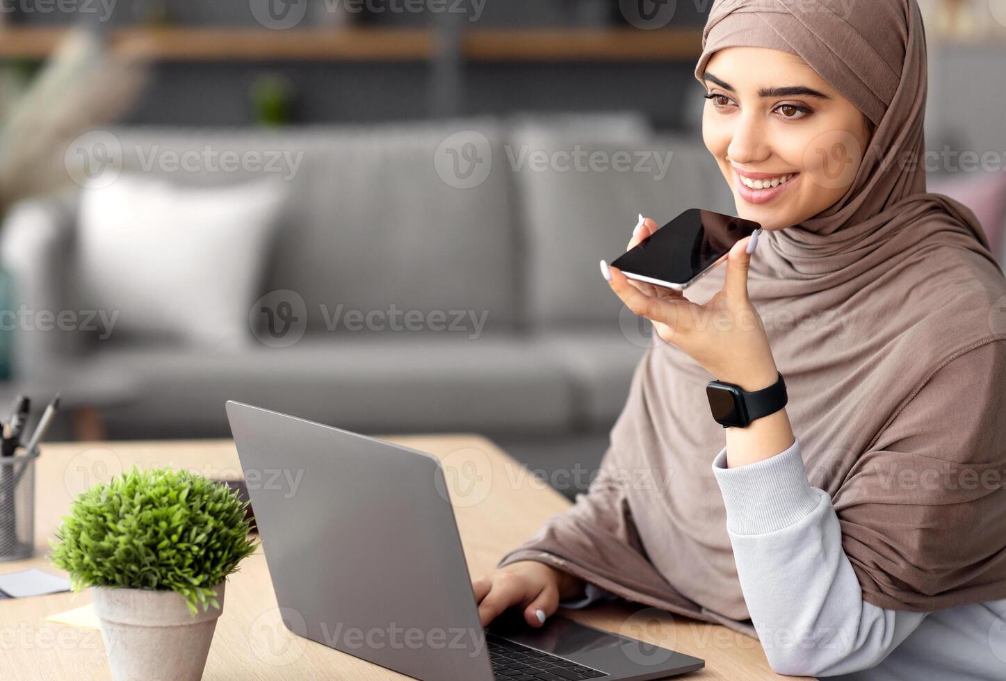 Work From Home. Young Muslim Business Lady Using Voice Search Assistant, Talking On Mobile Phone And Using Laptop Computer. Smiling Woman Sitting At Desk With Pc At Home Office, Wearing Hijab photo