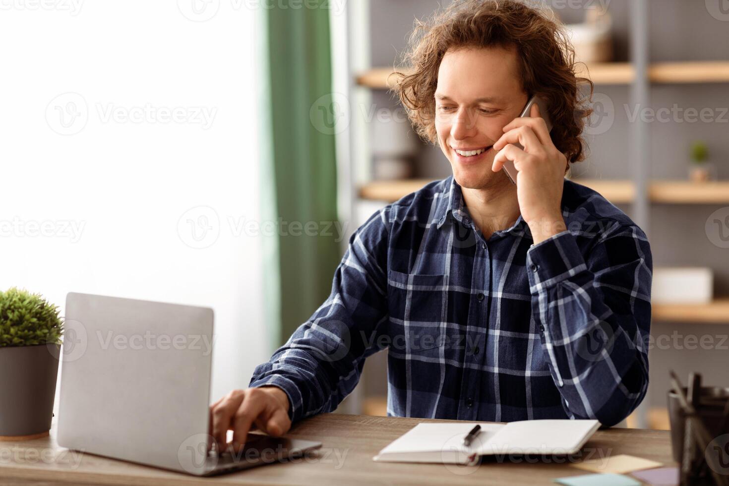 Cheerful Millennial Businessman Talking On Cellphone Working Online On Laptop Sitting At Workplace In Modern Office. Freelancer Man Chatting On Phone Indoors. Mobile Communication. Side View photo