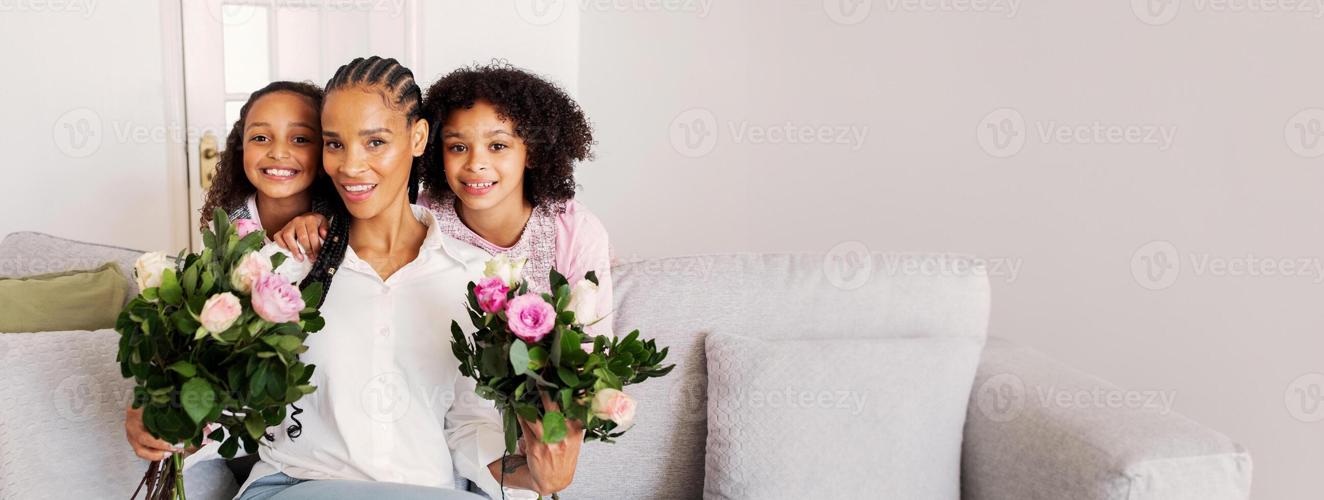 A black mother smiles while holding flower bouquets with her two daughters. They are sitting on a comfortable couch in a bright and inviting living room, sharing a joyful moment together. photo
