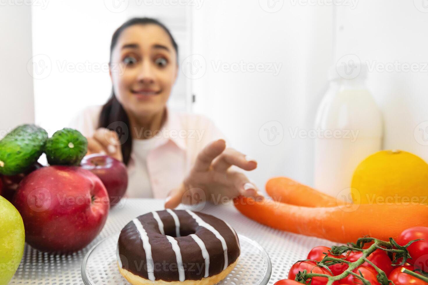 Junk Food And Cheat Meal Concept. Excited funny woman reaching and taking yummy donut from refrigerator at home, view from inside the fridge, lady tempted to eat dessert, selective focus photo
