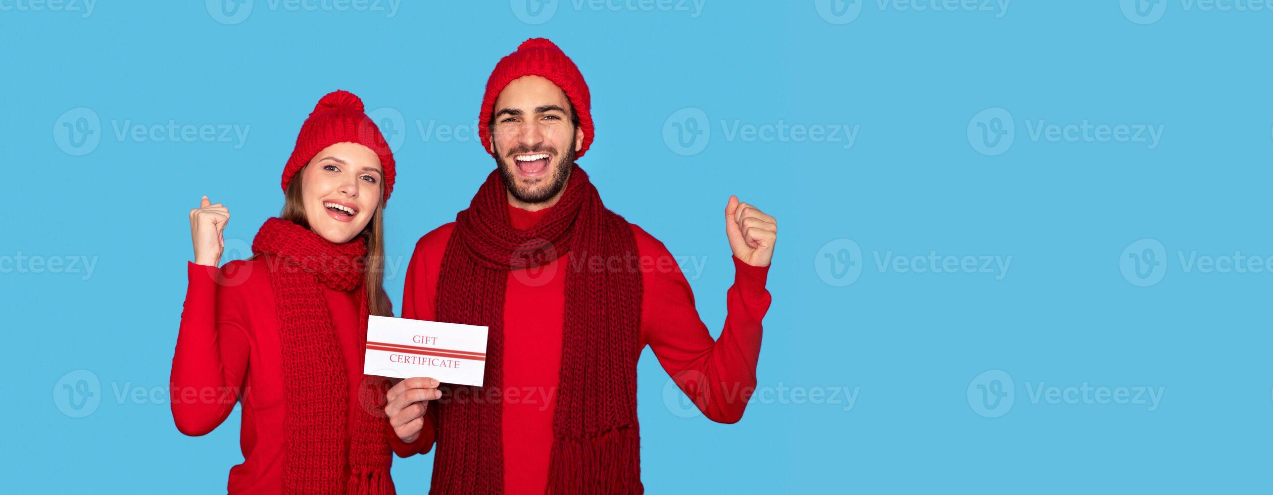 Joyful Young Couple In Knitted Hats Holding Gift Certificate In Hands And Celebrating Success, Happy Man And Woman Demonstrating Coupon Voucher And Shaking Fists, Copy Space photo