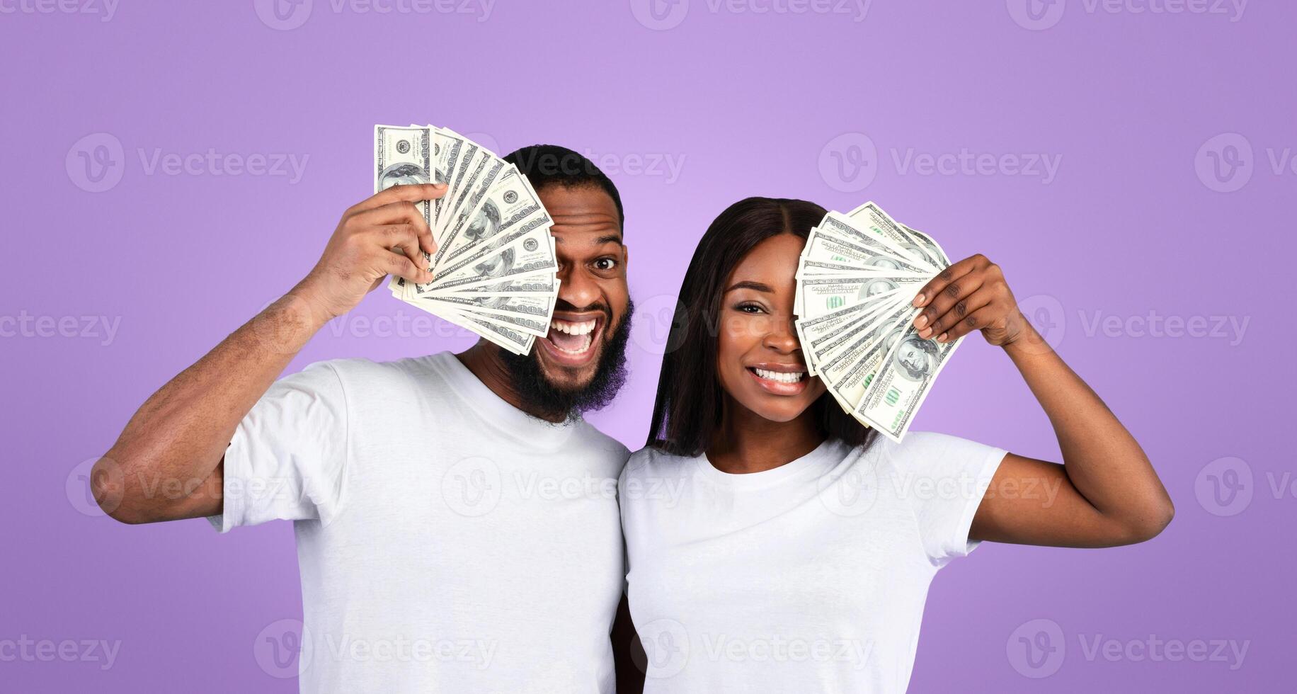 Big Win Concept. Portrait of excited young African American couple holding and covering eyes with a lot of dollar cash fan, celebrating success together, posing with money over blue studio background photo
