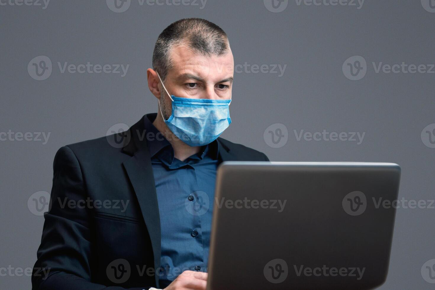Entrepreneur Man Working Using Laptop Computer Wearing Protective Mask Sitting On Gray Background. Distant Work Concept. Studio Shot photo