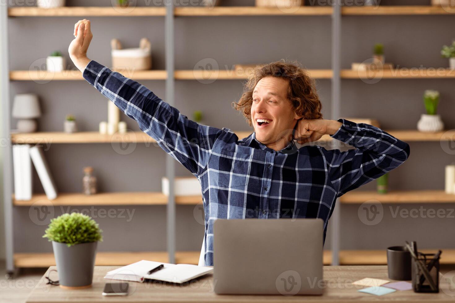 Funny Freelancer Guy At Laptop Stretching Arms And Yawning After Work On Computer Sitting At Desk In Modern Office. Successful Millennial Male Relaxing And Resting At Workplace Indoors photo
