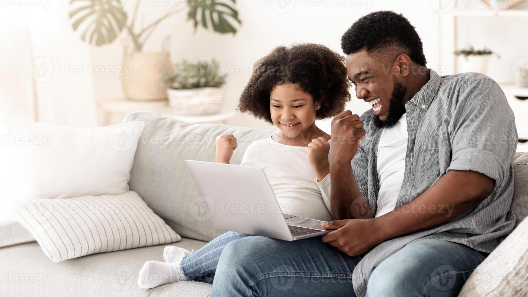 Excited black father and little daughter playing computer games on laptop at home, celebrating win with clenched fists, sitting on couch, copy space photo