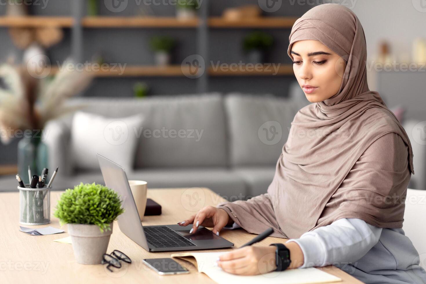 Home Office And Telecommuting Concept. Potrait of focused serious muslim woman in headscarf sitting at desk, using laptop. Female writing in notebook, taking notes, working remotely, free space photo