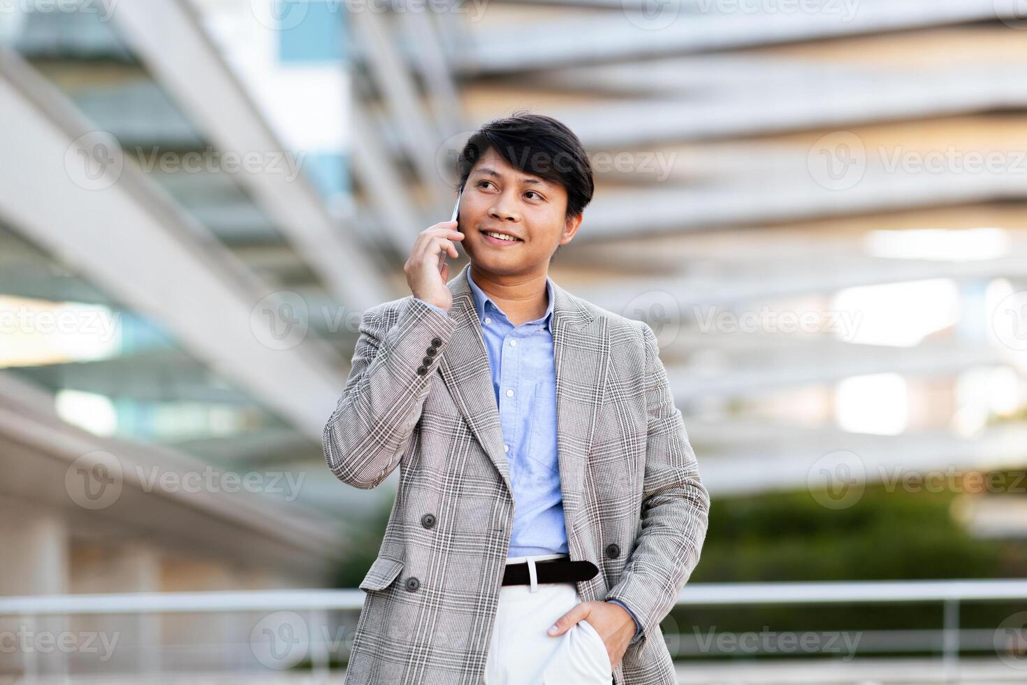 A young Asian man dressed in a fashionable blazer enjoys a phone conversation while standing outside a contemporary building in an urban setting, exuding confidence and style. photo