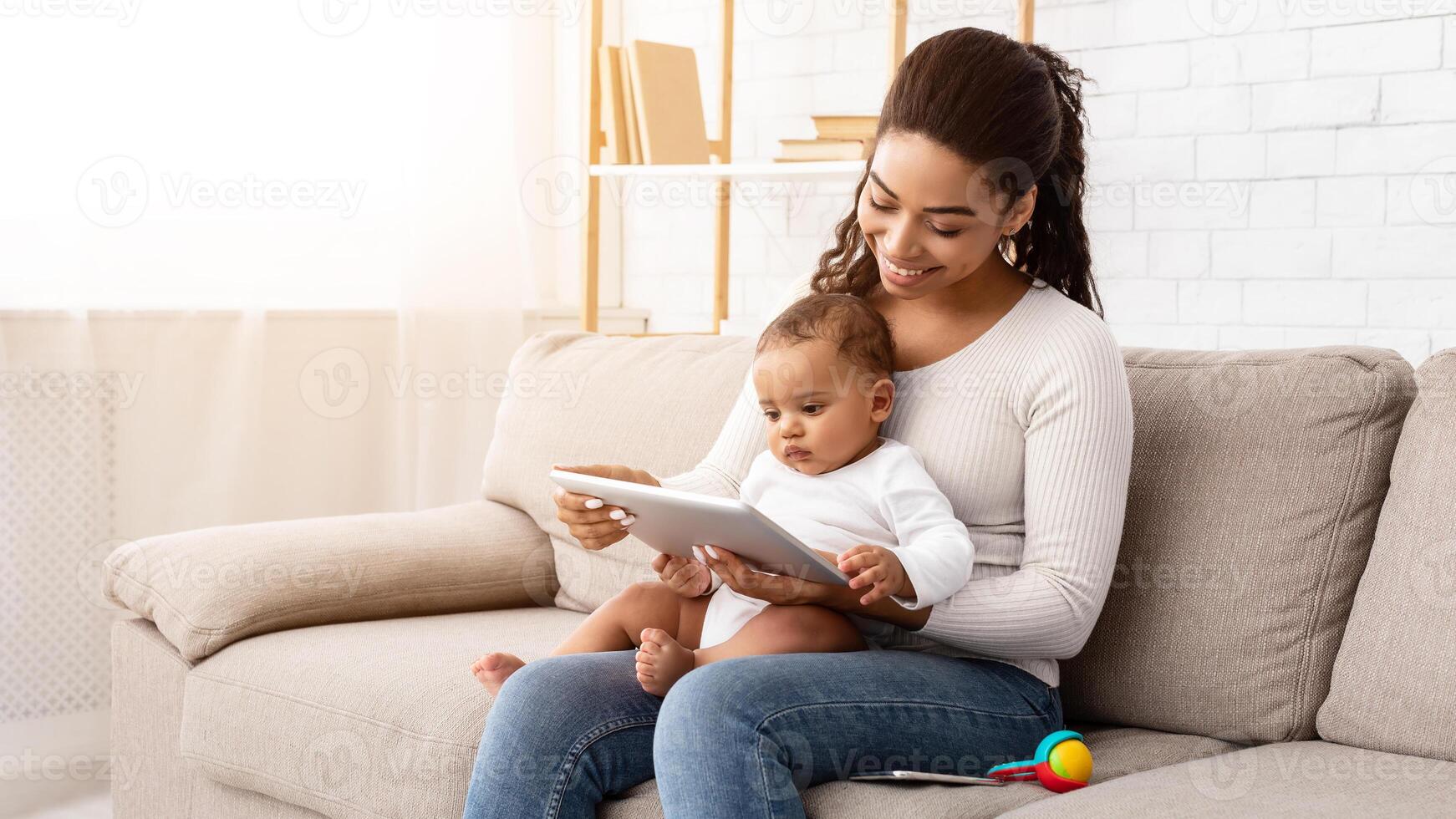 Cheerful Black Mommy Using Digital Tablet Holding Little Baby Showing Cartoons Online Sitting On Sofa In Living Room At Home. Mother Browsing Internet On Computer Using Games Application For Infant photo