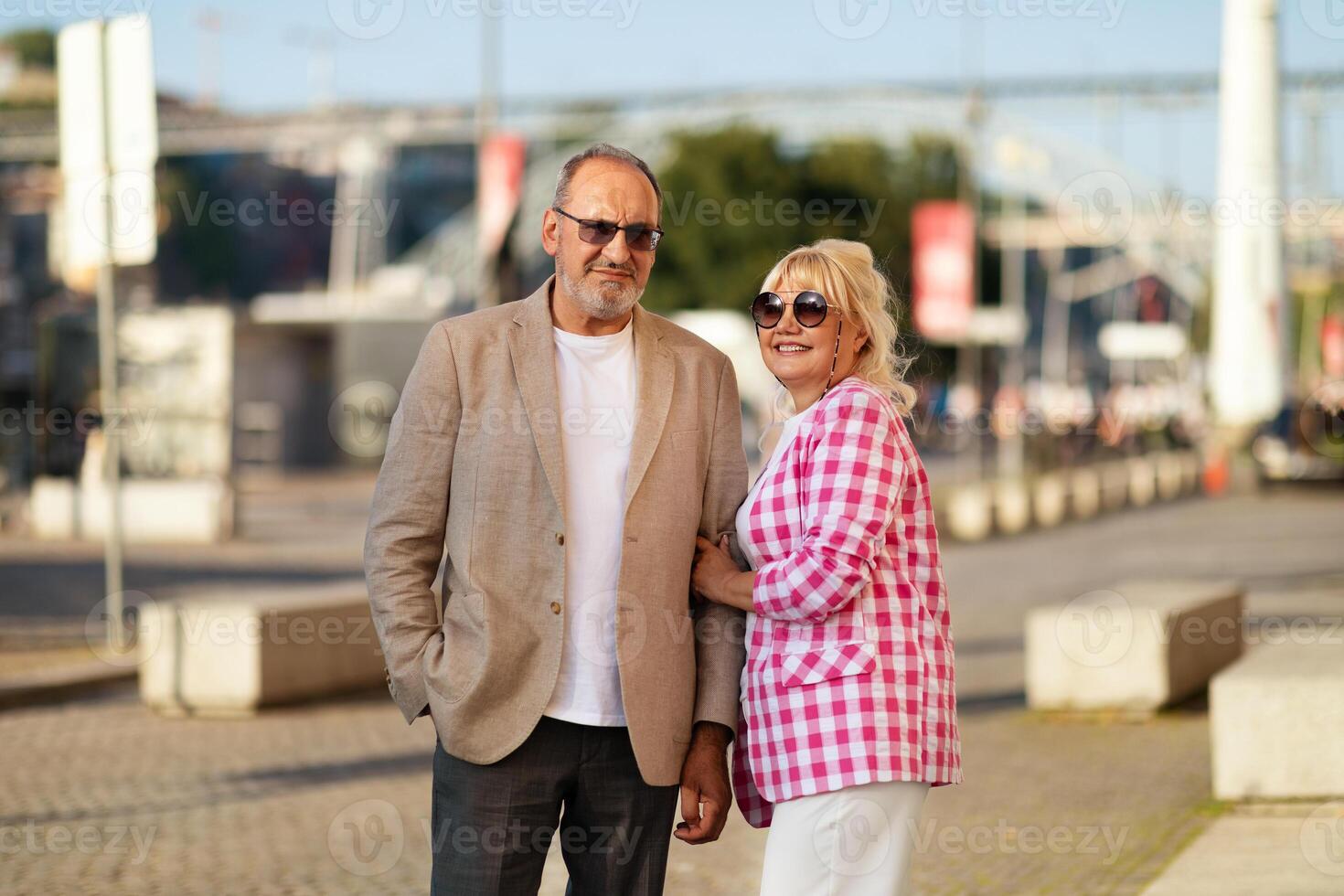 A man and woman stand side by side in a vibrant urban setting, both smiling. The woman wears sunglasses and a checked top while the man dons a blazer, creating a stylish appearance. photo