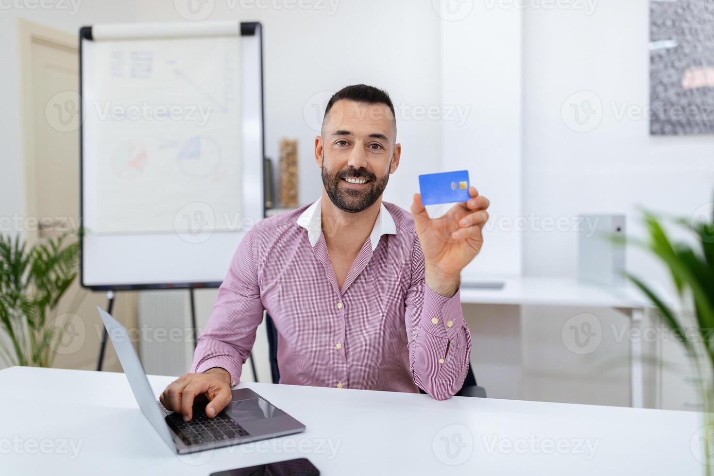 A man smiles as he holds up a credit card while sitting at a desk with a laptop open. The office features a whiteboard and green plants, creating a professional atmosphere. photo