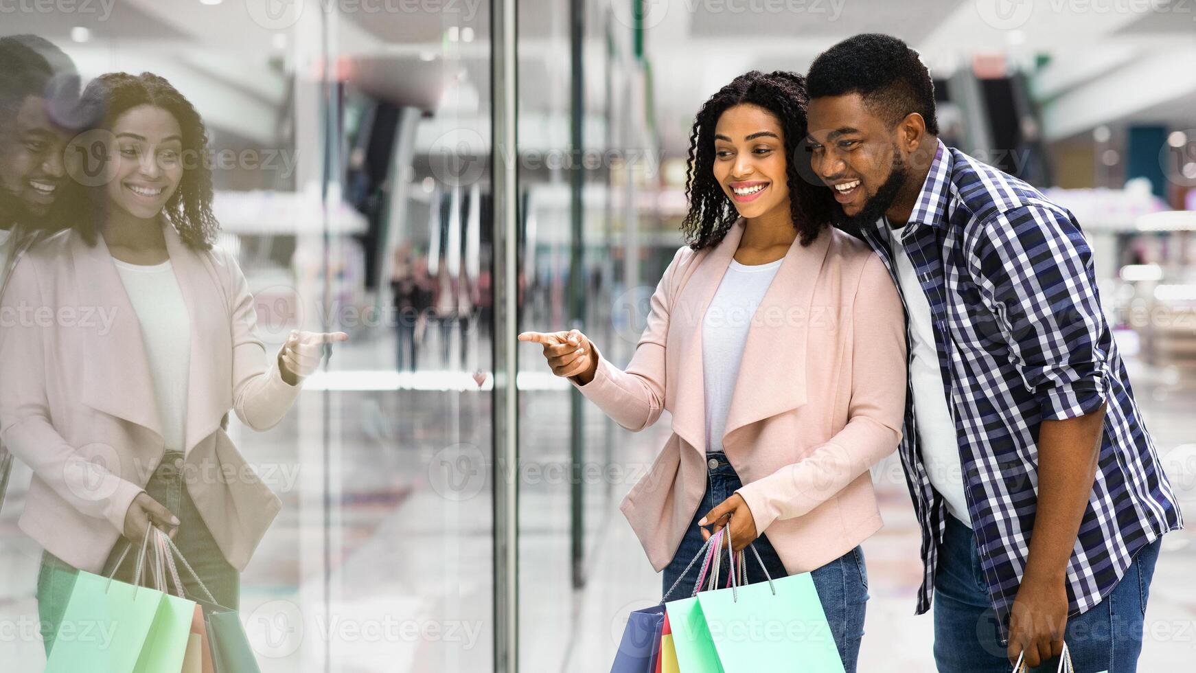 Happy Black Spouses Enjoying Shopping Together In Mall, Looking And Pointing At Showcase In Department Store, African American Couple Carrying Bright Shooper Bags With Purchases, Copy Space photo